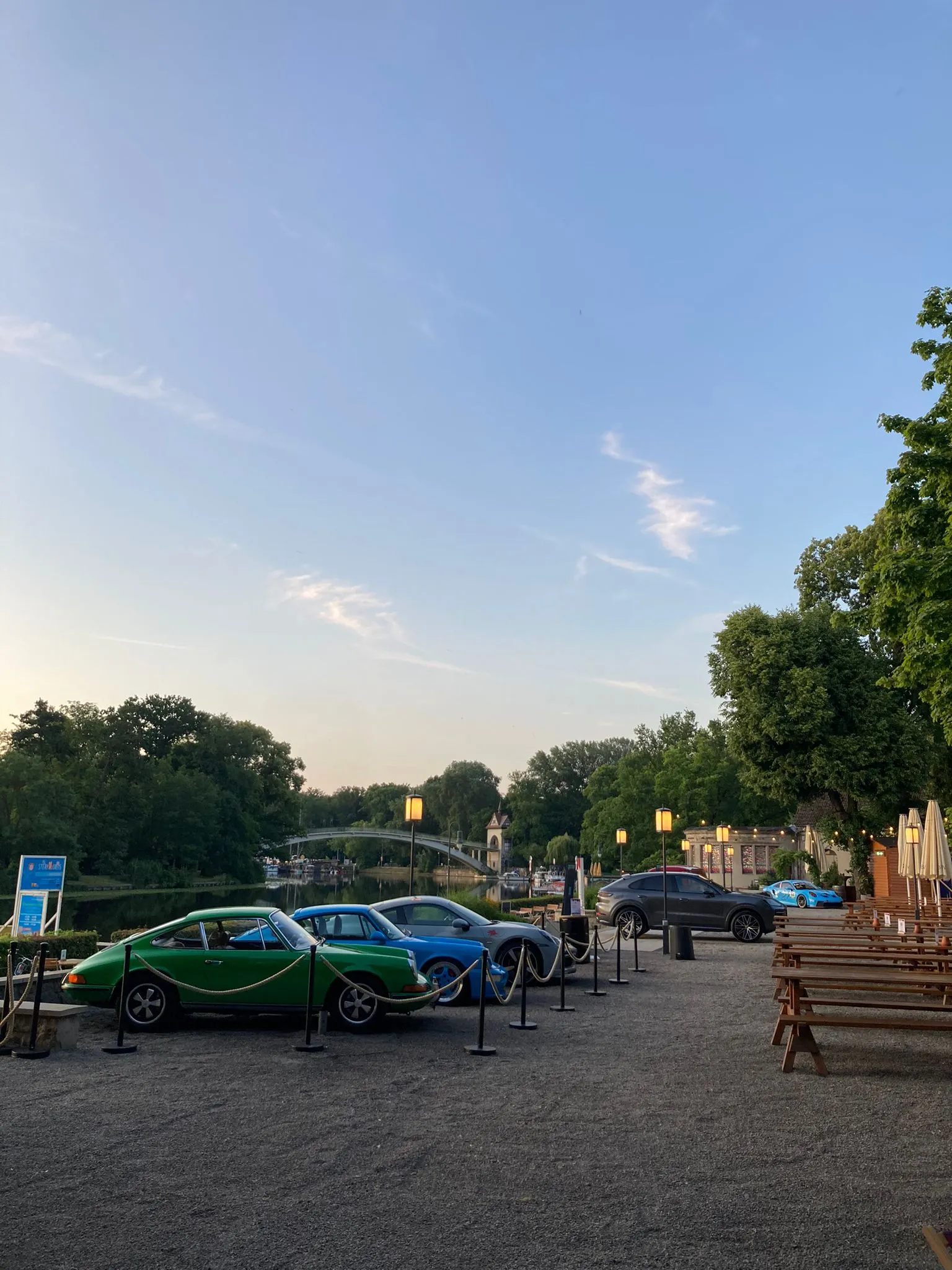 Oldtimer-Ausstellung am Flussufer in der Abenddämmerung: klassische Porsche hinter Kordeln, Biergarten-Bänke und eine Brücke im Hintergrund