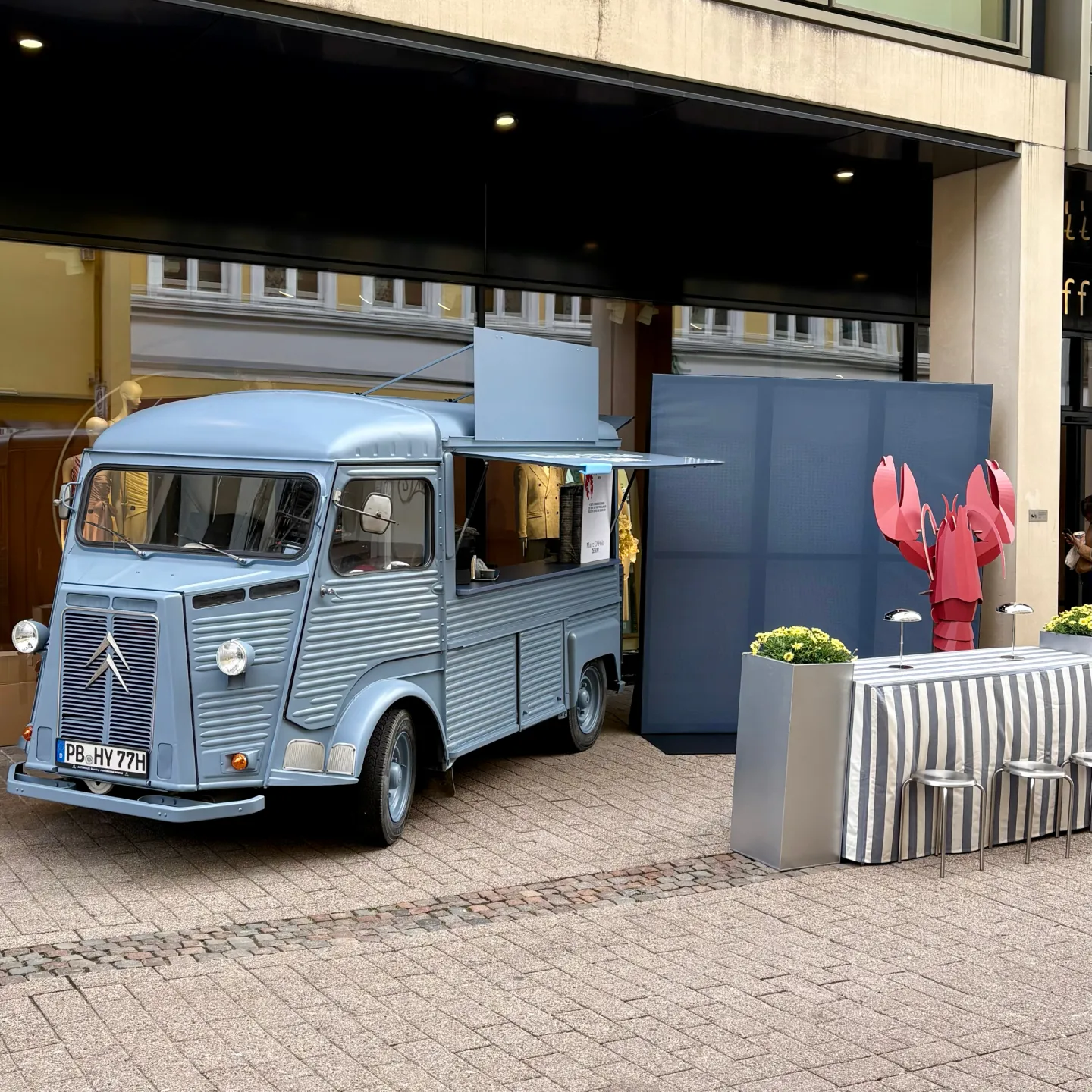 Hellblauer Citroën-HY-Foodtruck mit aufgeklappter Verkaufsklappe vor einem Kaufhaus, daneben gestreifte Stehtische und eine rote Hummer-Figur