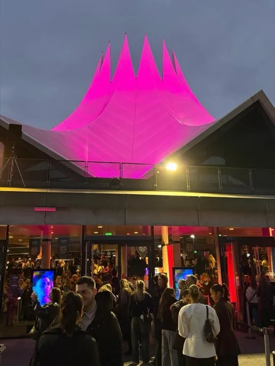 Pink angestrahltes Veranstaltungshaus, davor ankommende Gäste