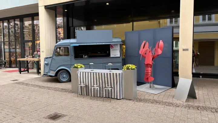 Roadshow-Setup mit gestreiften Stehtischen und roter Hummer-Figur vor dem Truck